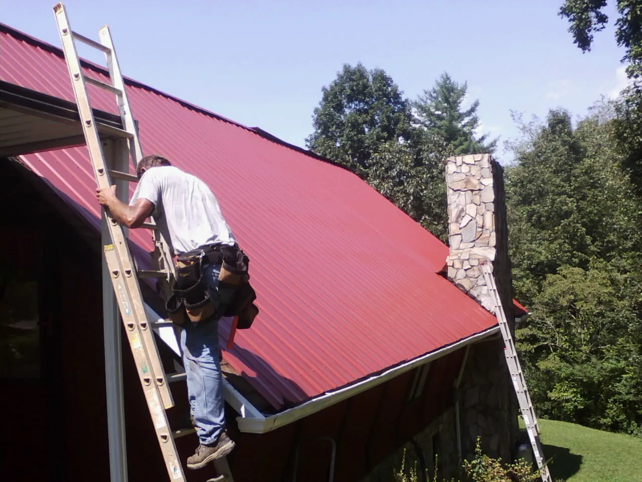 Roofer on a ladder beside a red metal roof and stone chimney — Soffit & Fascia Repair by Roofing Brothers in Loyalhanna